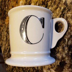 May include: White ceramic mug with a black letter "C" design. The mug has a wide base and a curved handle. The letter "C" is stylized with decorative elements. The mug is likely used for coffee or tea.