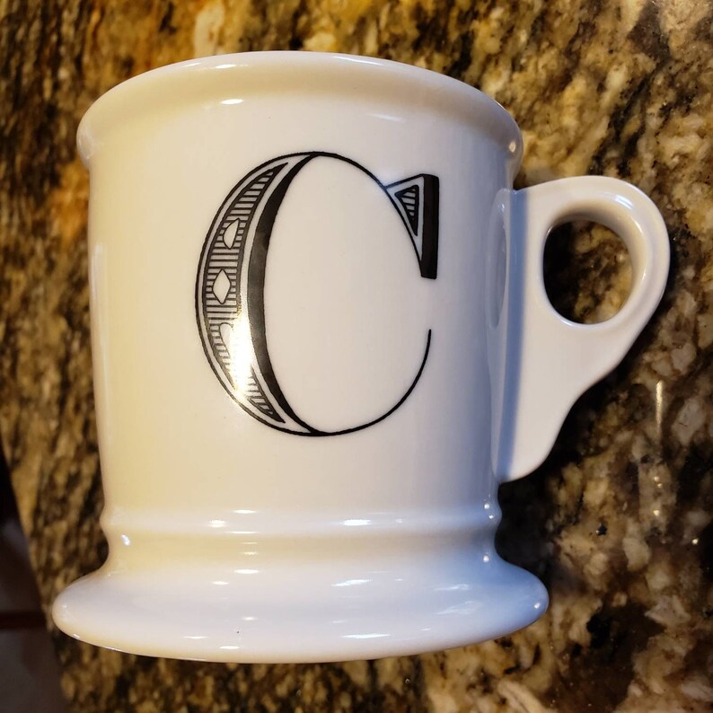 May include: White ceramic mug with a black letter "C" design. The mug has a wide base and a curved handle. The letter "C" is stylized with decorative elements. The mug is likely used for coffee or tea.