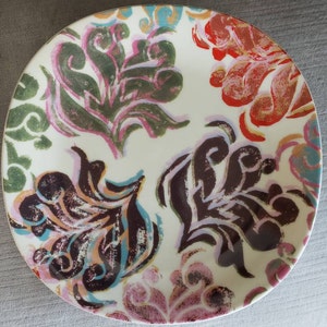 May include: A round, off-white ceramic plate with a colorful floral pattern. The design features stylized flowers in shades of green, red, brown, and pink, with accents of blue and orange. The plate has a smooth surface and a decorative edge.
