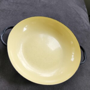 May include: A yellow enamel cast iron pan with black handles. The pan has a rounded shape and is sitting on a gray surface. The interior of the pan is a light yellow color.