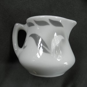 May include: A small, white ceramic creamer with a curved handle and spout. The creamer features a gray leaf pattern around the body. The creamer is designed for serving cream or milk.
