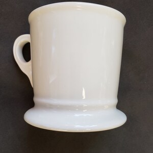 May include: A white ceramic mug with a handle. The mug has a flared base and a slightly flared rim. The mug is a solid, glossy white color. The handle is a simple loop shape.