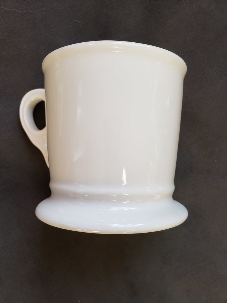 May include: A white ceramic mug with a handle. The mug has a flared base and a slightly flared rim. The mug is a solid, glossy white color. The handle is a simple loop shape.