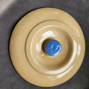 May include: A round, tan ceramic lid with a raised center and a blue knob. The lid has a glossy finish and appears to be for a jar or container. The background is a solid gray color.