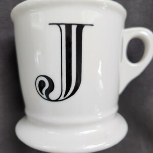 May include: White ceramic mug with a black letter "J" on the front. The mug has a handle and a flared base. The letter "J" is in a bold, stylized font.