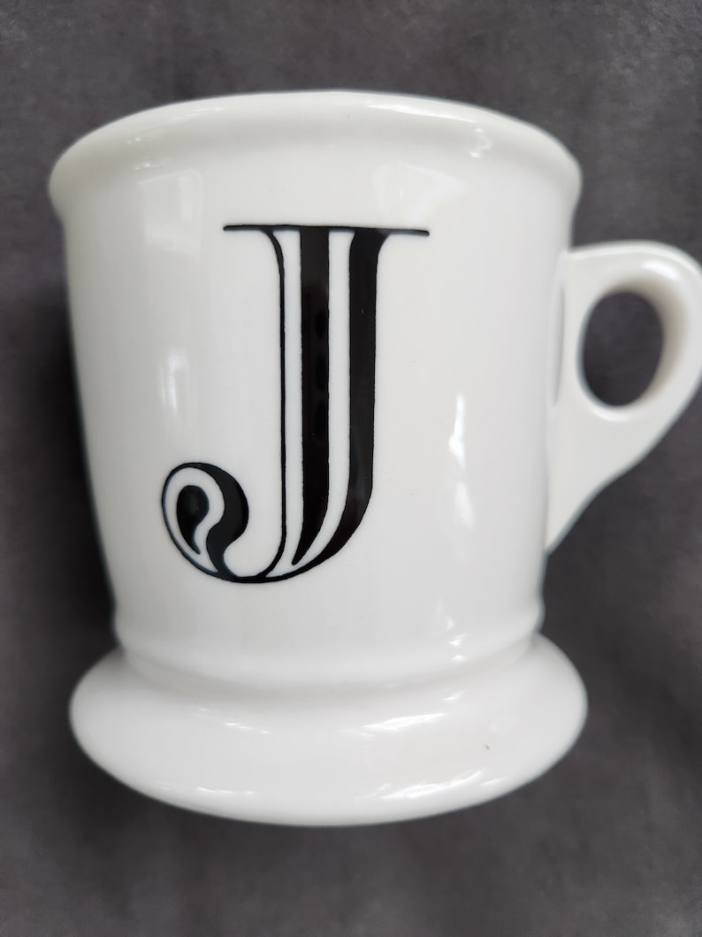 May include: White ceramic mug with a black letter "J" on the front. The mug has a handle and a flared base. The letter "J" is in a bold, stylized font.