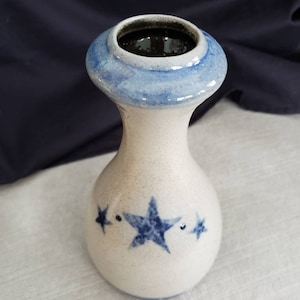 May include: A white ceramic vase with a blue rim and star designs. The vase has a bulbous base and a narrow neck that flares out at the top. The stars are dark blue and are scattered around the vase.