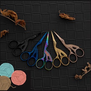 May include: Set of six decorative scissors with bird-shaped handles. The scissors are in various colours, including black, blue, gold, and rainbow. The scissors are arranged in a row on a black textured surface.