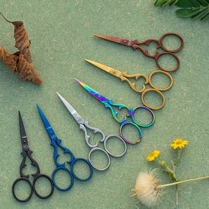 May include: Seven pairs of decorative scissors with ornate handles in a rainbow of colours, including black, blue, silver, rainbow, gold, and copper, arranged in a semi-circle on a green surface.