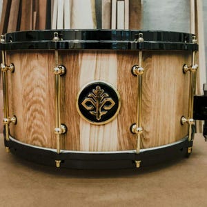 Puede incluir: Caja de madera con acabado de veta de madera natural, aros negros y herrajes dorados. Un emblema negro y dorado está centrado en el cuerpo del tambor. La caja está colocada sobre un fondo de tablones de madera.