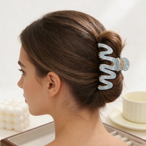 以下が含まれることがあります： 水色と白の大理石模様のヘアクリップ。クリップは波状のデザインで、茶色の髪のお団子をまとめています。クリップは光沢のある素材でできており、金色のトリムが付いています。