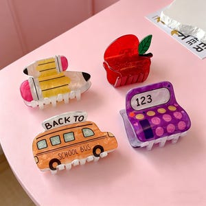 Op de afbeelding: Vier acryl haarclips in schoolthema-ontwerpen. Inclusief een potlood, een rode appel, een schoolbus met de tekst "BACK TO SCHOOL BUS" en een rekenmachine met "123" op het scherm. De clips zijn in verschillende kleuren, waaronder geel, oranje en paars.