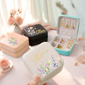 May include: A collection of small, square jewelry boxes in various colors, including pink, black, and light blue. Each box features floral designs and personalized names. One box is open, displaying rings and earrings.