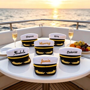 May include: Six white and black captain hats with gold braided trim and gold embroidered laurel leaves on the brim. Each hat features a unique name embroidered on the front: Sharon, Rita, Murray, Arabela, Dolores, and Jamie in various fonts.