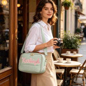 May include: A light green rectangular crossbody bag with a brown top handle and a wide matching shoulder strap. The front of the bag features the name Karleigh embroidered in pink script lettering. The bag has gold-toned metal zippers.