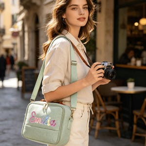 May include: A light green rectangular crossbody bag with a long adjustable shoulder strap. The front of the bag features the name "Emilia" embroidered in a colorful, gradient script font. The bag has a top zipper closure and gold hardware.