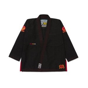 Pode incluir: Kimono de Jiu-Jitsu preto com acabamentos e costuras vermelhas. O kimono apresenta logotipos bordados nas mangas e na parte inferior da frente, e uma etiqueta com texto. Projetado para treinamento e competição de artes marciais.