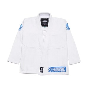 Puede incluir: Uniforme blanco de artes marciales con detalles azules. La chaqueta presenta un diseño envolvente, un cuello y el nombre de la marca "SHOOTOL" en azul en la parte inferior delantera. Hay logotipos circulares azules en los hombros. Una etiqueta negra es visible.