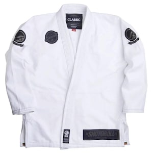 Pode incluir: Um uniforme de artes marciais branco, ou gi, com patches e texto pretos. O gi apresenta um design clássico com gola em V e costuras reforçadas. O nome da marca "SHOYOROLL" é exibido na parte inferior frontal.