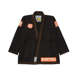 Puede incluir: Gi de Jiu-Jitsu negro con costuras y parches naranjas. El gi presenta el nombre de la marca "SHOYOROLL" en múltiples lugares. El gi tiene un cuello tradicional y está diseñado para el entrenamiento y la competición de artes marciales.
