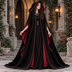 May include: A floor-length black hooded cloak with a deep red lining and intricate floral embroidery along the edges. The garment features a large buckle at the neck and three smaller buckles down the front, with wide open sleeves and a full skirt.