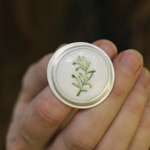 May include: A silver ring with a white porcelain top featuring a green botanical design.