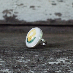 May include: A silver ring with a white ceramic top featuring a hand-painted yellow lemon with green leaves.