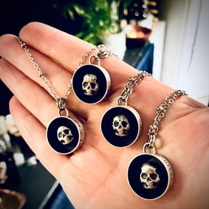 May include: Four silver necklaces with black circular pendants featuring detailed skull designs. Each pendant has a black background and a silver skull with intricate details.