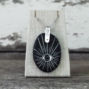 May include: A black ceramic pendant featuring a white eye design with radiating lines. The pendant is attached to a silver chain with a silver leaf-shaped bail.