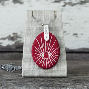 May include: A red oval pendant necklace with a white eye and sunburst design. The pendant is attached to a silver chain with a silver charm featuring a leaf design.