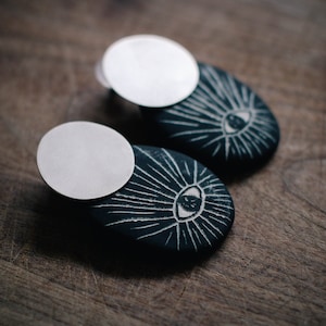 May include: A pair of black and white eye-shaped earrings with a silver disc on top. The earrings are made of clay and have a textured surface.