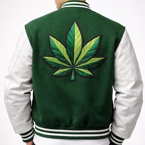 Op de afbeelding: Groene en witte varsity jas met een groot, geborduurd cannabisbladontwerp op de rug. De jas heeft witte mouwen, een groene body en gestreepte details op de manchetten en zoom. Een stijlvol bovenkledingstuk.