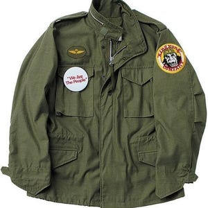 Puede incluir: Chaqueta de estilo militar verde oliva con cierre de cremallera, múltiples bolsillos y cuello alto. Cuenta con un pequeño parche con un emblema alado y un botón con el texto "We Are The People". Un parche de "King Kong Company" está en la manga derecha.