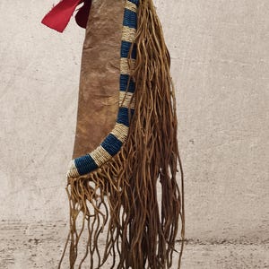 Puede incluir: Una funda de cuero marrón con un diseño de cuentas en azul y crema, y ​​largas y finas franjas de cuero marrón. Una pieza de tela roja está unida cerca de la parte superior, y un objeto oscuro en forma de cuerno está en la parte superior.