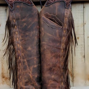 May include: Brown leather chaps with decorative floral tooling on the yoke. The chaps have a fringed edge and a pocket detail. The leather has a distressed appearance, and the chaps are hanging against a wooden background.