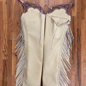 May include: Cream-colored leather chaps with intricate floral tooling on the upper portion. The chaps feature a pocket, fringe along the sides, and a belt strap. The leather is a light tan color, and the fringe has blue accents.