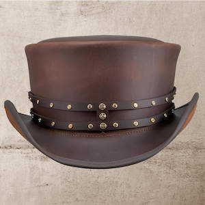 May include: A brown leather top hat with a wide brim. The hat features a dark brown leather band around the crown, adorned with small, gold-colored studs. The hat's design is reminiscent of vintage or steampunk styles.