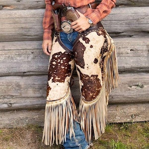 May include: Cowhide chaps with long fringe, worn over blue jeans. The chaps have a brown and white spotted pattern. A brown leather holster with a gun is visible. The person is wearing a plaid shirt and cowboy boots.
