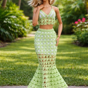 May include: A doll in a light green and white crochet two-piece outfit. The top is a halter neck style, and the skirt is a mermaid style. The doll has blonde hair and wears white heels.