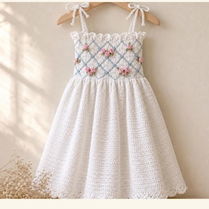 May include: A white crochet sundress featuring a smocked bodice with a blue lattice pattern and small pink rosebud embellishments. The dress has thin tie-shoulder straps and a scalloped hem. Text reads Stitching Villa Rosebud Lattice Smocked Sundress.