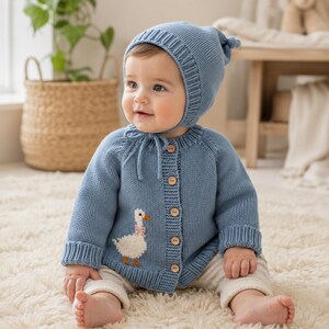 May include: A dusty blue knitted cardigan and matching bonnet set. The cardigan features a white goose appliqué with a pink bow, long sleeves with ribbed cuffs, and five light brown wooden buttons down the front. The bonnet has a ribbed edge.