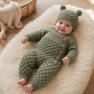 Può includere: Una tutina in maglia per neonati color verde salvia con un motivo a rilievo testurizzato sulle gambe e sul busto, maniche lunghe e chiusura laterale con piccoli bottoni in legno. Il completo include un berretto abbinato con due pon pon.