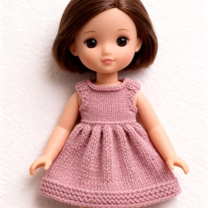 Puede incluir: Muñeca con cabello y ojos marrones que lleva un vestido rosa tejido. El vestido tiene un escote redondo y una falda plisada. La muñeca está sobre un fondo blanco.