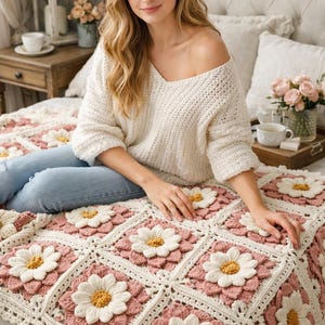 May include: A handmade crochet blanket featuring a daisy flower design. The blanket has a white background with pink and white daisy flowers. The blanket is on a bed with a person wearing a white sweater and blue jeans.