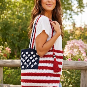May include: A rectangular crochet tote bag featuring a red, white, and blue American flag pattern. The bag has a dark blue section with white stars, horizontal red and white stripes, two shoulder straps, and a red tassel with wooden beads.