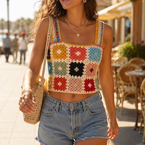May include: A sleeveless crop top made of multi-colored crochet granny squares in shades of yellow, red, blue, pink, and black, featuring a square neckline and thick straps, paired with light blue denim shorts with a frayed hem and a straw bag.