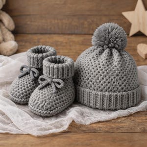 以下が含まれることがあります： グレーのニットベビーセット。リボン付きのベビーブーツのペアと、ポンポンが付いたお揃いの帽子が含まれています。セットは、木製の表面に白い布の上に置かれています。