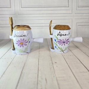 Peut inclure: Deux pots en céramique blanche avec couvercles et cuillères en bois. Chaque pot a une poignée et un motif floral avec les mots "Cornichons" et "Vignare". Les pots sont sur une surface en bois clair.