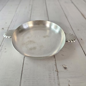 Peut inclure: Un plat rond en métal argenté avec deux poignées décoratives. Le plat a une cuvette peu profonde et une surface légèrement réfléchissante. Les poignées sont ornées d'un motif en forme d'éventail. Le plat est posé sur un fond en bois blanc.