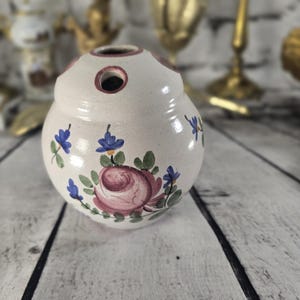 Peut inclure: Un vase à fleurs en céramique crème avec un corps rond et plusieurs trous sur le dessus. Le vase est décoré d'une rose rose et d'accents floraux bleus. Le vase repose sur une surface en bois.